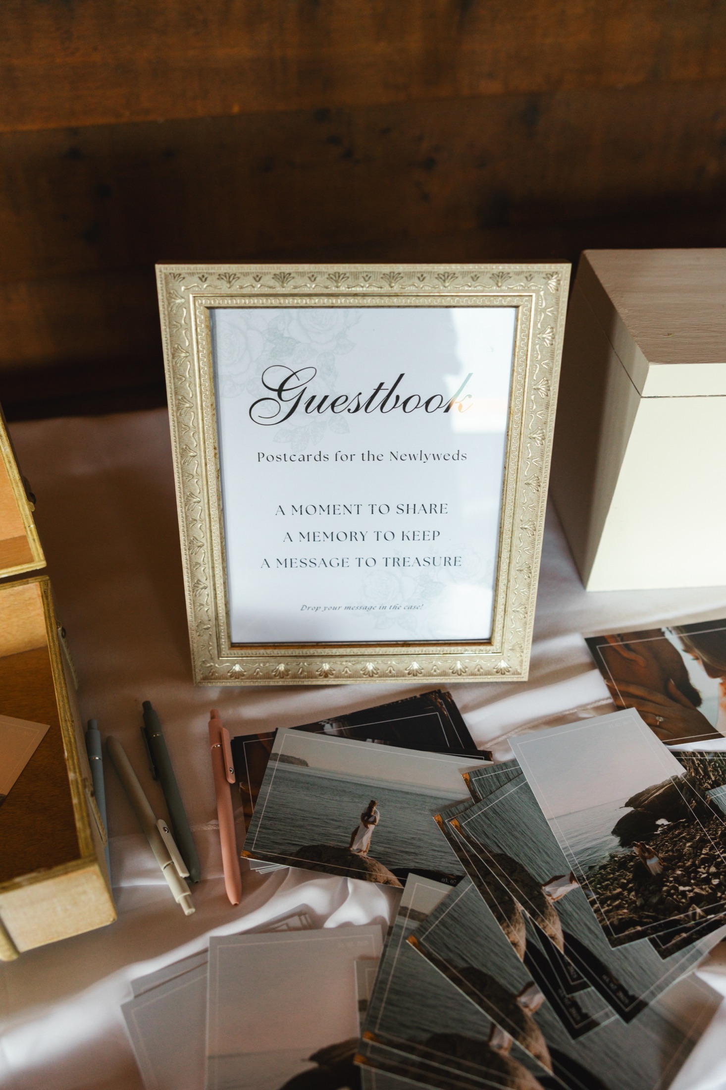 A framed guestbook sign in a gilt frame beside fanned postcards and a row of coloured pens on a pale cloth.