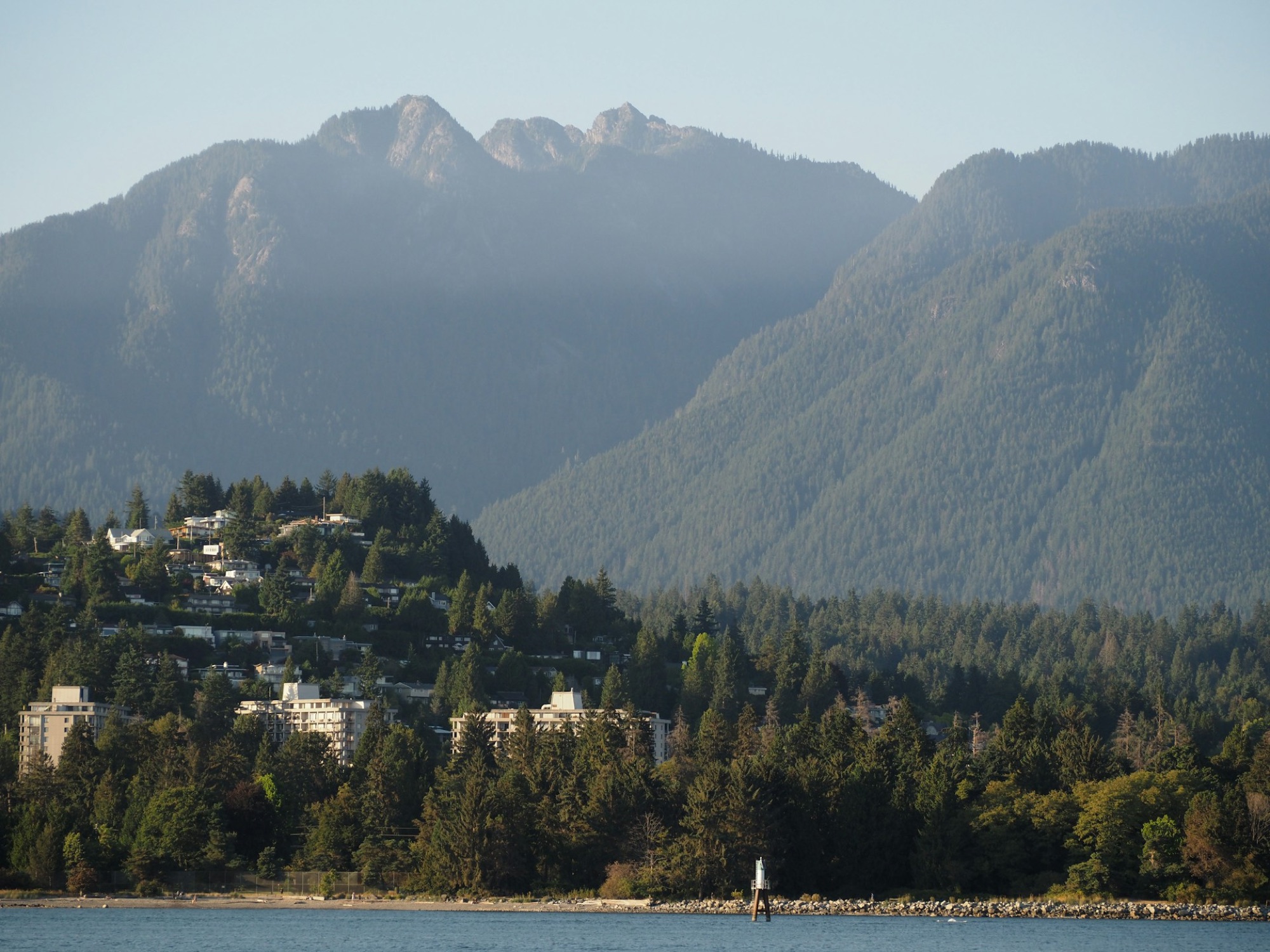 Forested mountains rising above a wooded shoreline of houses, seen across open water in soft afternoon haze.